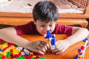 Asian boy is getting creative with assembling colorful plastic bricks into robots and planes on a wooden table happy and fun at home.Kid Creators concept. photo