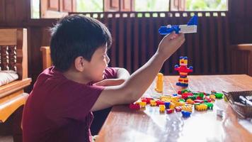 Asian boy is getting creative with assembling colorful plastic bricks into robots and planes on a wooden table happy and fun at home.Kid Creators concept. photo