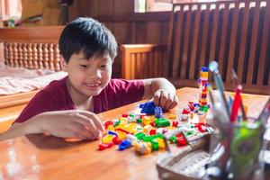 Asian boy is getting creative with assembling colorful plastic bricks into robots and planes on a wooden table happy and fun at home.Kid Creators concept. photo