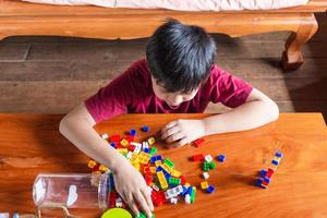 Asian boy is getting creative with assembling colorful plastic bricks into robots and planes on a wooden table happy and fun at home.Kid Creators concept. photo