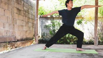 concepto de vida saludable de un joven asiático que practica yoga asana pose de guerrero de pie, ejercicio y poses en una alfombra de yoga verde. ejercicio al aire libre en el jardín. estilo de vida saludable. foto