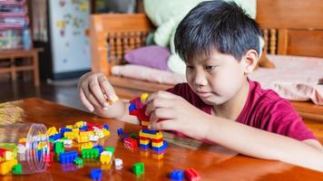 Asian boy is getting creative with assembling colorful plastic bricks into robots and planes on a wooden table happy and fun at home.Kid Creators concept. photo