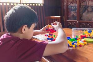Asian boy is getting creative with assembling colorful plastic bricks into robots and planes on a wooden table happy and fun at home.Kid Creators concept. photo