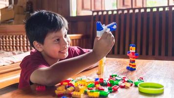 Asian boy is getting creative with assembling colorful plastic bricks into robots and planes on a wooden table happy and fun at home.Kid Creators concept. photo