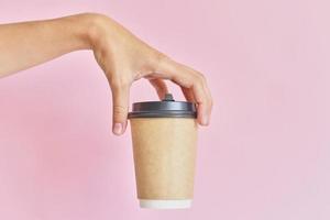 Female hand holding paper cup for coffee take away on pink background photo