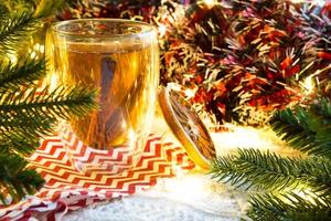 Transparent double-walled glass tumbler with hot tea and cinnamon sticks on the table with Christmas decor. New year's atmosphere, slice of dried orange, garland and tinsel, spruce branch, cozy photo