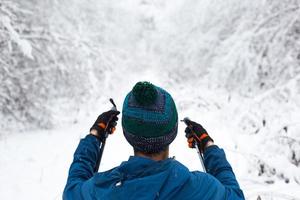 Skier in windbreaker and hat with pompom with ski poles in his hands with his back against the background of a snowy forest. Cross-country skiing in winter forest, outdoor sports, healthy lifestyle. photo