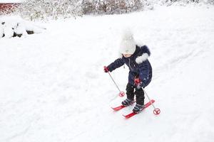 Children's feet in red plastic skis with sticks go through the snow from a slide-a winter sport, family entertainment in the open air. A little girl glides down the slope from an early age. Copy space photo
