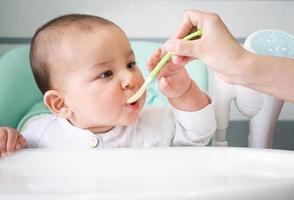 Mom feeds the baby with a spoon of vegetable puree at the children's feeding table. Baby's appetite, healthy nutrition, introduction of complementary foods. Copyspace, mock up photo