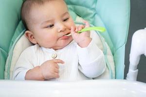 A hungry baby is gnawing on a plastic spoon at the table on a high chair. Teething, whims, itchy gums, introduction of complementary foods photo