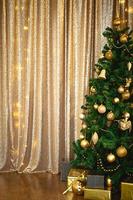 Christmas green artificial fir tree in the Golden room with lights garlands decorated with Golden balls, beads and toys. The box with the gifts in foil and black paper. New Year. Space for text photo