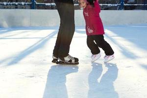 papá le enseña a su pequeña hija a patinar sobre hielo en una pista de patinaje en el patio de edificios de varios pisos en la ciudad. día soleado de invierno helado, deportes de invierno activos y estilo de vida foto