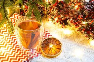 Transparent double-walled glass tumbler with hot tea and cinnamon sticks on the table with Christmas decor. New year's atmosphere, slice of dried orange, garland and tinsel, spruce branch, cozy photo