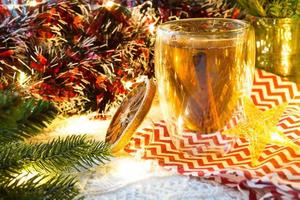 Transparent double-walled glass tumbler with hot tea and cinnamon sticks on the table with Christmas decor. New year's atmosphere, slice of dried orange, garland and tinsel, spruce branch, cozy photo