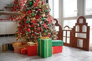 el árbol de navidad está decorado con juguetes rojos y verdes, cajas con regalos en el suelo. ambiente de año nuevo en la casa, interior festivo. foto