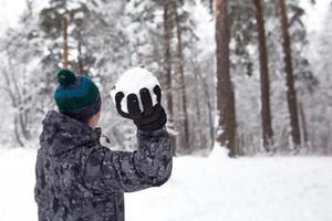 un joven con una bola de nieve en la mano se está divirtiendo, balanceándose para lanzar. juegos familiares y amistosos de invierno y entretenimiento en el bosque con nieve al aire libre foto