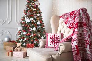 Christmas tree with red and white decor in a white living room with gifts in boxes, a chair with pillows and a blanket with winter ornaments, a fireplace, a fur rug. New year, European style. photo