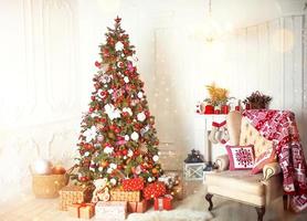 Christmas tree with red and white decor in a white living room with gifts in boxes, a chair with pillows and a blanket with winter ornaments, a fireplace, a fur rug. New year, European style. photo