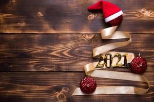 A Christmas tree made of gold ribbon with the numbers 2021 is decorated with red balloons on a dark wooden background. Flat lay. Space for text. New year, Santa hat. photo