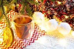 Transparent double-walled glass tumbler with hot tea and cinnamon sticks on the table with Christmas decor. New year's atmosphere, slice of dried orange, garland and tinsel, spruce branch, cozy photo