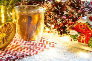 Transparent double-walled glass tumbler with hot tea and cinnamon sticks on the table with Christmas decor and small house. New year's atmosphere, slice of dried orange, garland, spruce branch, cozy photo