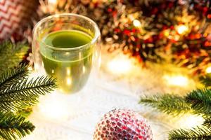 vaso transparente de vidrio de doble pared con té matcha japonés en la mesa con decoración navideña. atmósfera de año nuevo, guirnaldas y oropel, rama de abeto, acogedora manta de punto, pelota, servilleta a rayas