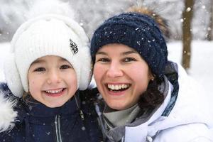 Mom and daughter in warm clothes in the snowy forest hugging and looking into the frame. Winter entertainment outside, active recreation, fun in the cold . Childhood, strong family. Portrait photo