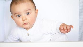 Cute portrait of a baby in a crib close-up. A child in white clothes on white underwear. Tenderness and care, children's problems. photo