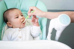 Mom feeds the baby with a spoon of vegetable puree at the children's feeding table. Baby's appetite, healthy nutrition, introduction of complementary foods. Copyspace, mock up photo