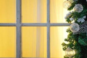 Christmas decorations made of spruce branches and white transparent balls and ribbons frame the window with wooden frame and yellow light. New year festive atmosphere, comfort of home. Space for text photo