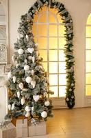 Christmas green artificial fir tree in a bright room of the house near a large floor-to-ceiling window with yellow light. Decorated with white balls, cones. Gift boxes in Kraft paper. New Year. photo