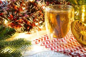 Transparent double-walled glass tumbler with hot tea and cinnamon sticks on the table with Christmas decor. New year's atmosphere, slice of dried orange, garland and tinsel, spruce branch, cozy photo