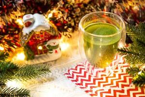 Transparent double-walled glass tumbler with Japanese matcha tea on table with Christmas decor. New year's atmosphere, garland and tinsel, spruce branch, cozy, knitted blanket, ball, striped napkin photo