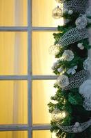 Christmas decorations made of spruce branches and white transparent balls and ribbons frame the window with wooden frame and yellow light. New year festive atmosphere, comfort of home. Space for text photo
