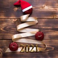 A Christmas tree made of gold ribbon with the numbers 2021 is decorated with red balloons on a dark wooden background. Flat lay. Space for text. New year, Santa hat. photo