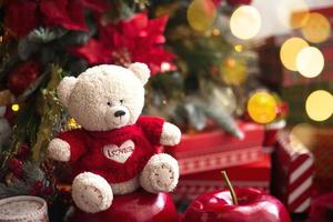 oso de peluche blanco con suéter de punto rojo con corazón en el pecho y las palabras amor cerca del árbol de navidad entre las cajas de regalo. regalo para tu amada, declaración de amor, decoración de poinsettia. copie el espacio foto