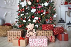 Gifts in boxes and a Teddy bear close-up under a Christmas tree with red and white decor in a white living room. New year, European style, kitchen decor. Space for text, selective focus photo