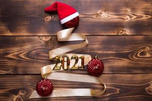 A Christmas tree made of gold ribbon with the numbers 2021 is decorated with red balloons on a dark wooden background. Flat lay. Space for text. New year, Santa hat. photo