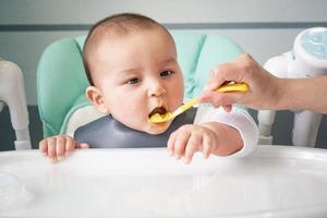 Mom feeds the baby with a spoon of vegetable puree at the children's feeding table. Baby's appetite, healthy nutrition, introduction of complementary foods. Copyspace, mock up photo