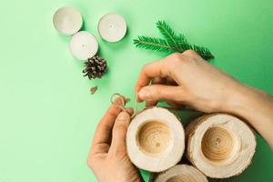 DIY Christmas candle holder made of pine logs, candles, craft rope, fir branches and cones. Hands fasten and tie the rope. Step-by-step instructions flat lay, step 3. photo