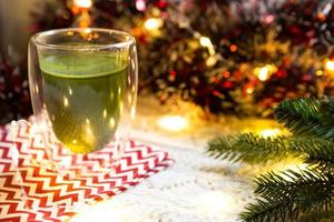 vaso transparente de vidrio de doble pared con té matcha japonés en la mesa con decoración navideña. atmósfera de año nuevo, guirnaldas y oropel, rama de abeto, acogedora manta de punto, pelota, servilleta a rayas