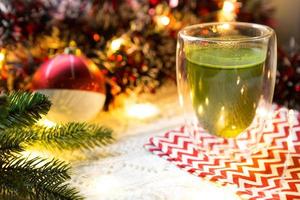 Transparent double-walled glass tumbler with Japanese matcha tea on table with Christmas decor. New year's atmosphere, garland and tinsel, spruce branch, cozy, knitted blanket, ball, striped napkin photo