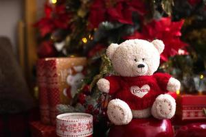 White Teddy bear in red knitted sweater with heart on the chest and the words Love near Christmas tree among the gift boxes. Gift to your beloved, Declaration of love, poinsettia decor. Copy space