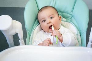 un bebé hambriento está mordiendo una cuchara de plástico en la mesa de una silla alta. dentición, caprichos, picor de encías, introducción de alimentos complementarios foto