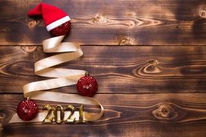 A Christmas tree made of gold ribbon with the numbers 2021 is decorated with red balloons on a dark wooden background. Flat lay. Space for text. New year, Santa hat. photo