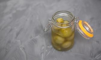 Pickled green small tomatoes in a glass jar with a lid on a gray background. Salted cherries, home-made, olives in brine. photo