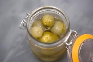 pequeños tomates verdes en escabeche en un frasco de vidrio con tapa sobre un fondo gris. cerezas saladas, caseras, aceitunas en salmuera. foto