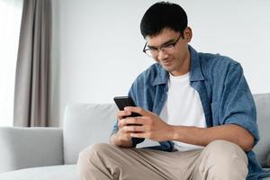 Happy Asian man using smartphone on sofa in living room at home, searching data and social media on internet. photo