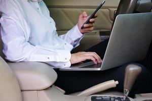Businessman working on laptop and mobile phone while sitting on driver seat in car. Lifestyle concept. photo