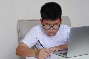 The boy with glasses sit on sofa is studying through laptop and writing in notebook during online lesson.  Education, learning and technology concept. photo